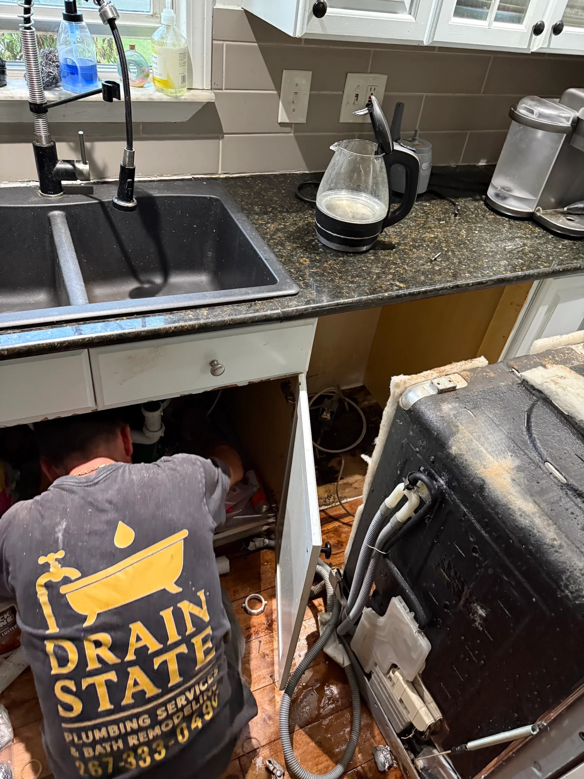 Plumber working under kitchen sink cabinet with granite countertop and black sink basin visible